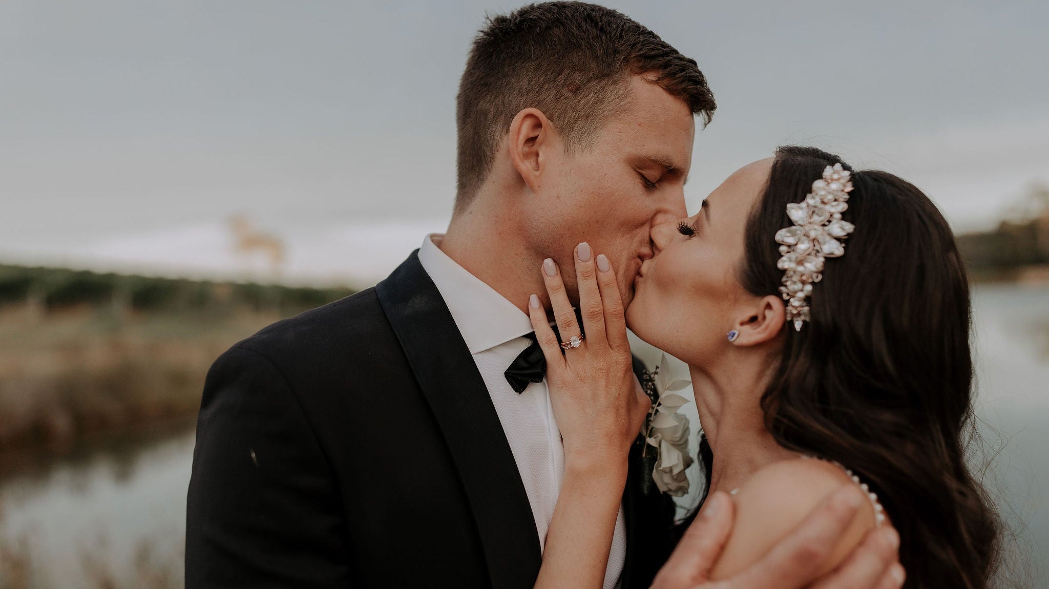 a stunning bride with dark hair wearing a crystal flower and pearl comb on the side of a sleek sides and glam wave hairstyle. Bride is kissing her groom