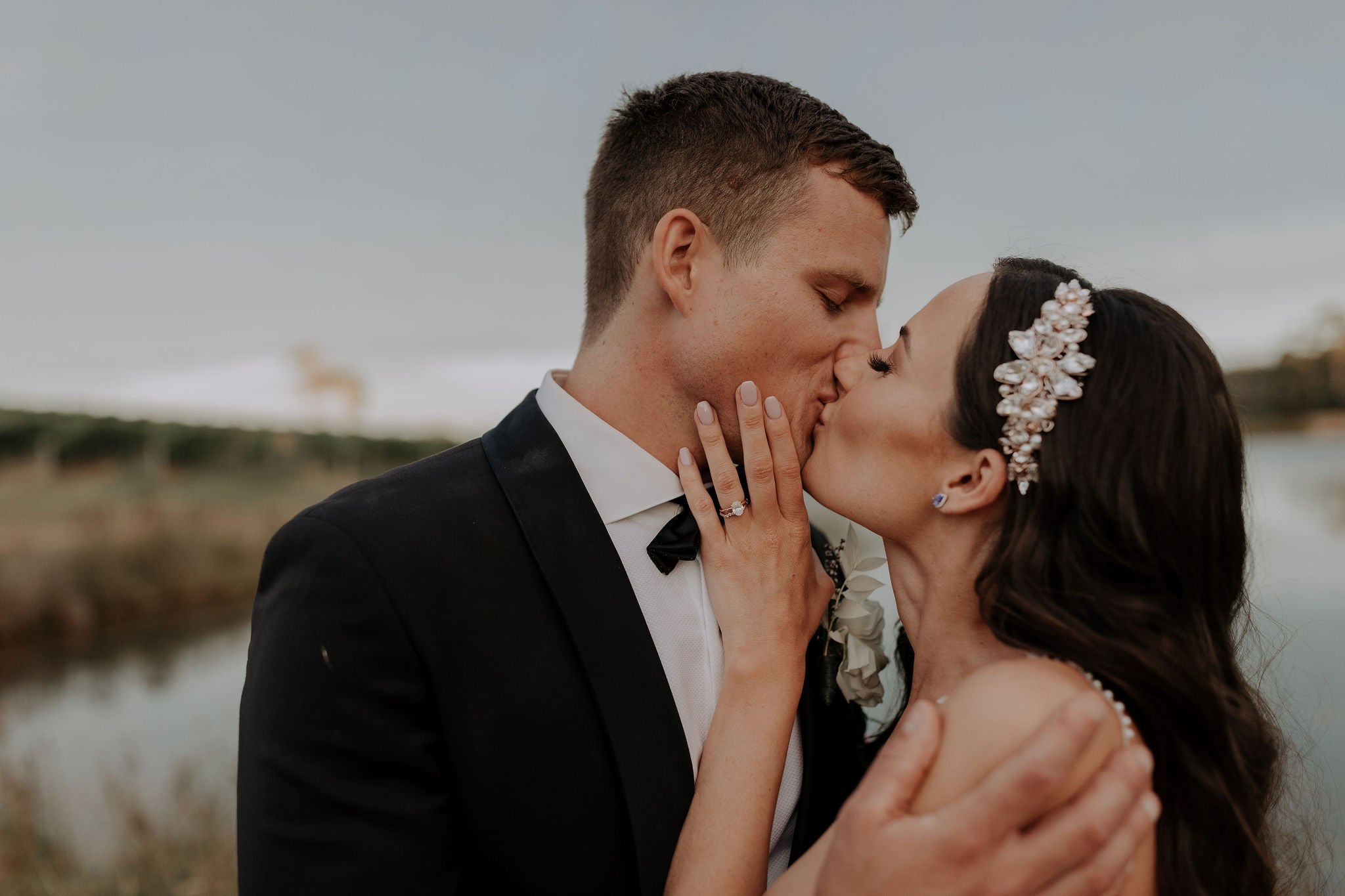 a stunning bride with dark hair wearing a crystal flower and pearl comb on the side of a sleek sides and glam wave hairstyle. Bride is kissing her groom