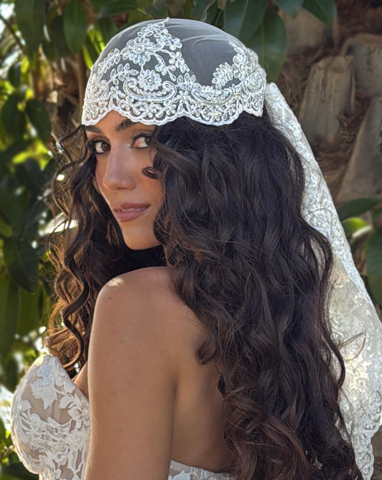 bride in tropical setting wearing a lace trim bandana veil like a cap. she is wearing a lace bodice. bride has long brown hair in beautiful curls