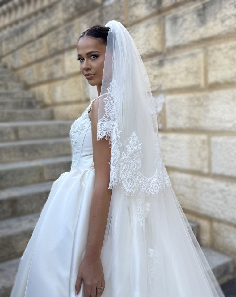 side view of a bride standing on stairs with a beautiful two tier full wedding veil that has a deep chantilly lace style trim around the border.