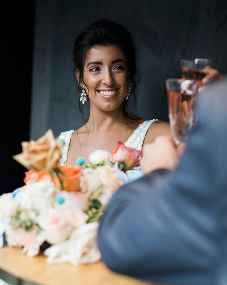 image of bride and groom sitting at table toasting champaign the bride is wearing earings white opal boho earing called pipa by kezani jewellery