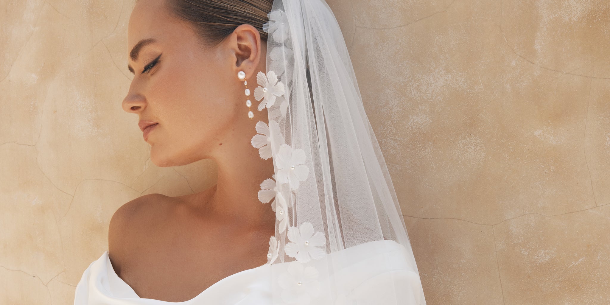 bride leaning on wall wearing her hair up with a long pearl multi drop with a d floral edge veil. off shoulder wedding gown - lola knight for kezani