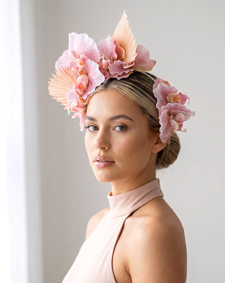 A woman wearing a large pink floral orchid fascinator headpiece with layered paper-like fan and a tall feather, paired with a dusky pink sleeveless top, photographed against a soft light gray background by kezani