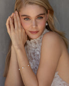 blonde hair model with hands up to her face. She is wearing a high collar lace blocked halter wedding gown and is wearing the faith dainty freshwater pearl bracelet and large pearl simple earrings