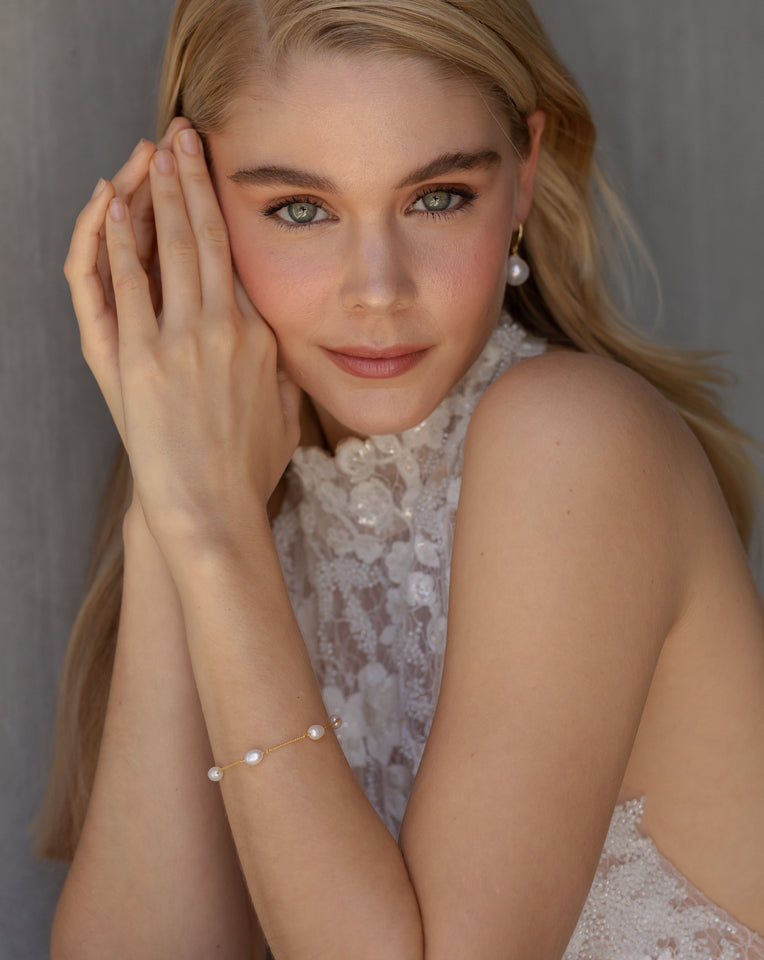 blonde hair model with hands up to her face. She is wearing a high collar lace blocked halter wedding gown and is wearing the faith dainty freshwater pearl bracelet and large pearl simple earrings