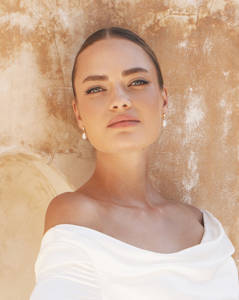 bride leaning on wall wearing gold dainty pearl earring with off shoukder gown - kezani jewellery